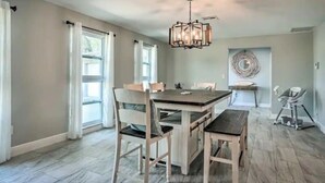 Un comedor con una mesa y sillas de madera, una araña y vistas al exterior a través de amplios ventanales.
