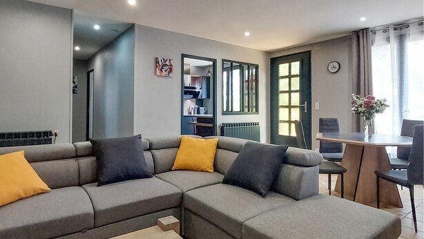 Un salon moderne avec un canapé d’angle gris, une table à manger avec des chaises et un coin cuisine visible à travers une porte ouverte.