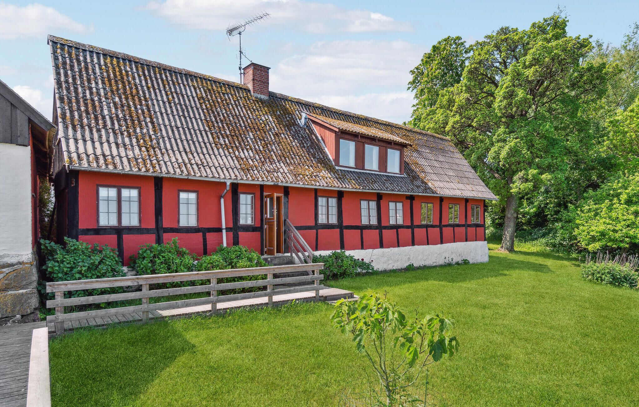 Une maison traditionnelle à colombages, aux couleurs rouge et blanche, avec un toit en pente, un escalier en bois menant à l’entrée et une pelouse bien entretenue.