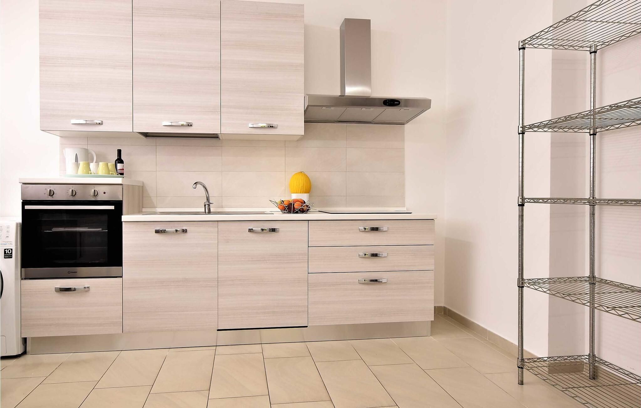 A modern kitchen with light wood cabinetry, a stainless steel range hood, a built-in oven, and a metal shelving unit.