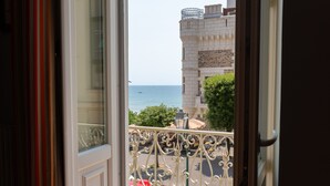 A balcony with a wrought iron railing, a red car parked in front, and a view of the sea and buildings in the background.