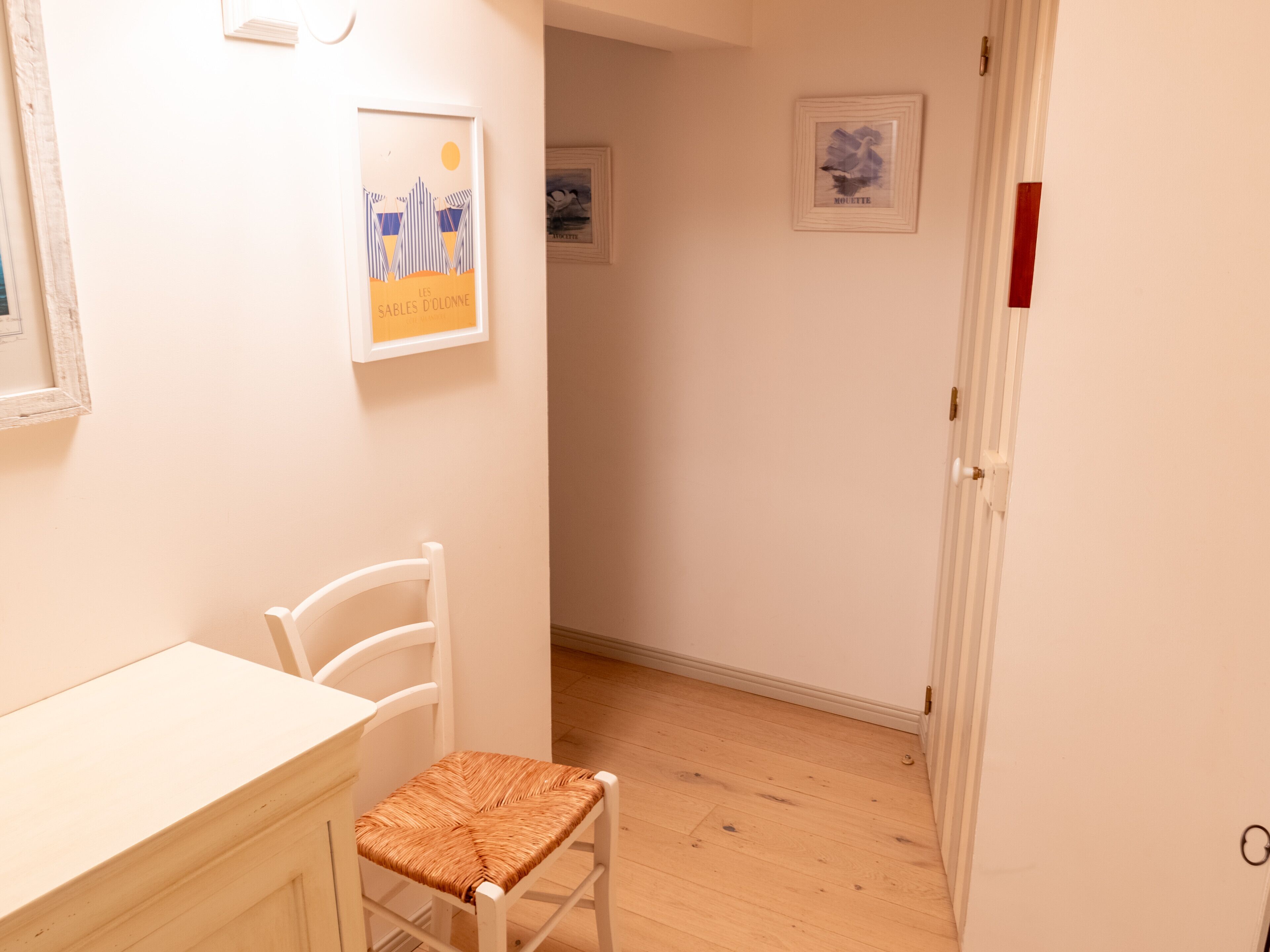 A narrow hallway with a white door on the right, a white chair with a woven seat, and a white table on the left.
