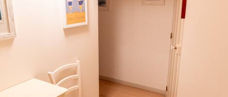 A narrow hallway with a white door on the right, a white chair with a woven seat, and a white table on the left.
