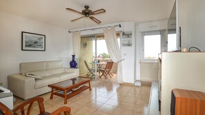 Una sala de estar con sofá, mesa de centro y sillas. Hay un ventilador de techo, una ventana con cortinas y una puerta que da a un área exterior.
