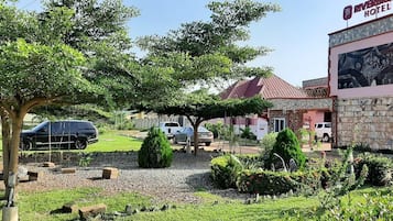 A hotel with a stone facade, a red roof, and a garden area with trees and shrubs.