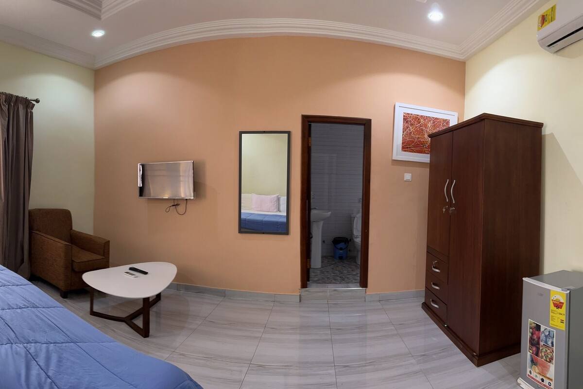 A hotel room with a bed, a small table, a chair, a wardrobe, and a bathroom visible through an open door.