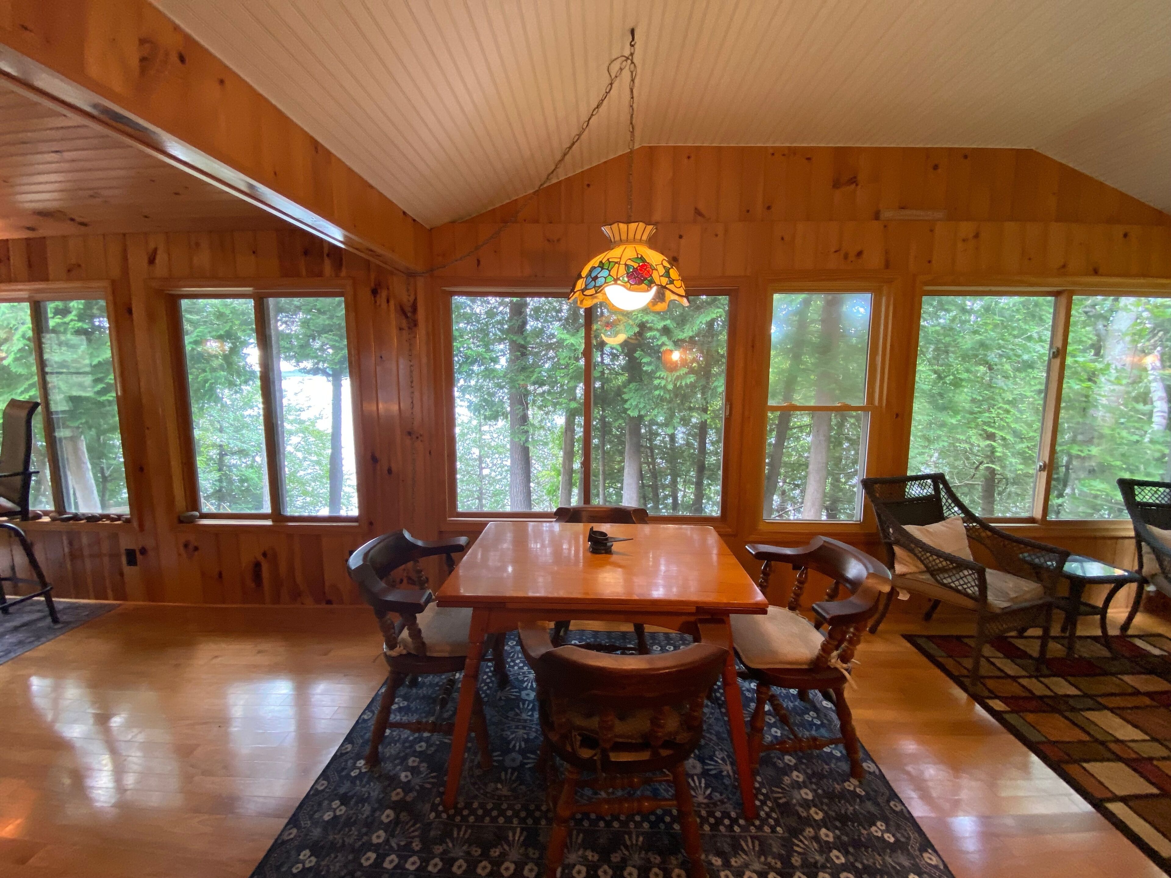 A wooden cabin with a dining table and chairs, large windows, and a hanging light fixture.