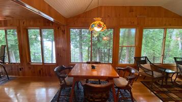 A wooden cabin with a dining table and chairs, large windows, and a hanging light fixture.