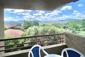 A balcony with a view of a mountainous landscape, a red-roofed building, and two blue and white chairs.