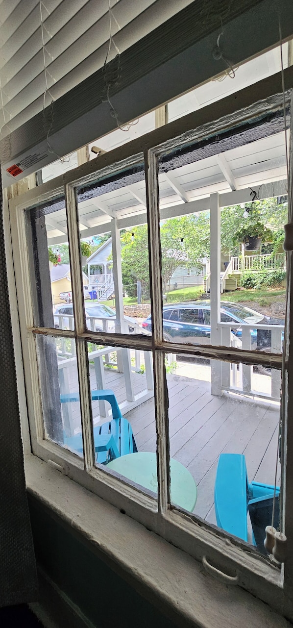 Blick durch ein Fenster mit weißem Rahmen, der einen überdachten Außenbereich mit blauen Stühlen und einem Tisch, ein Holzdeck und ein geparktes Auto zeigt.
