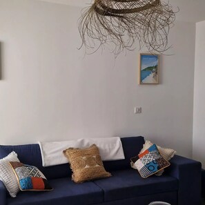 Un salón con un sofá azul oscuro, tres cojines estampados, una lámpara colgante tejida y un cuadro en la pared.