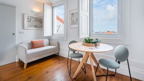 Una habitación con un sofá blanco, una mesa redonda de madera con sillas y dos ventanas con marcos blancos.
