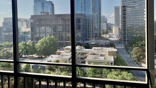 A cityscape through a window with multiple high-rise buildings, including a distinctive blue glass building. - ATL Downtown Condo w Balcony with City views (Atlanta)