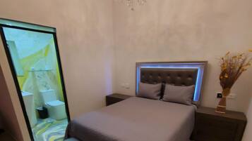 A bedroom with a bed, bench, nightstands, a chandelier, and a bathroom visible through a glass door.