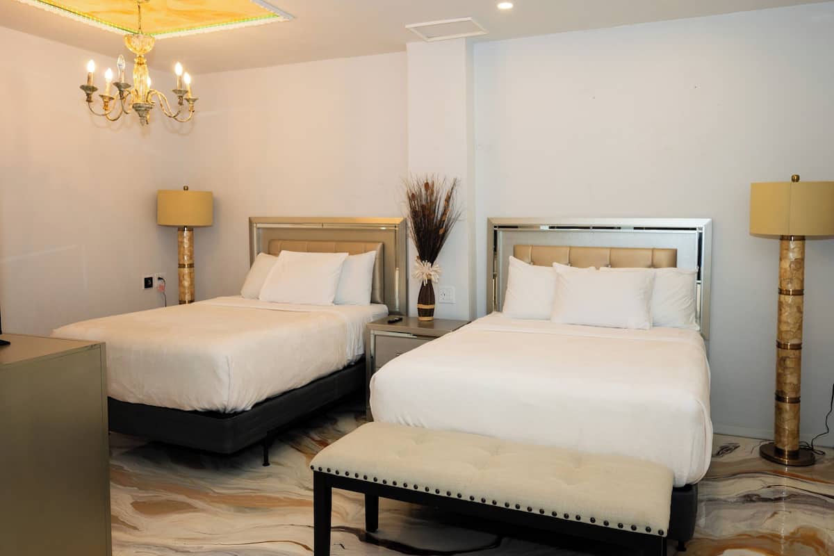 A hotel room with two beds, a bench, a chandelier, and a vase with dried plants.