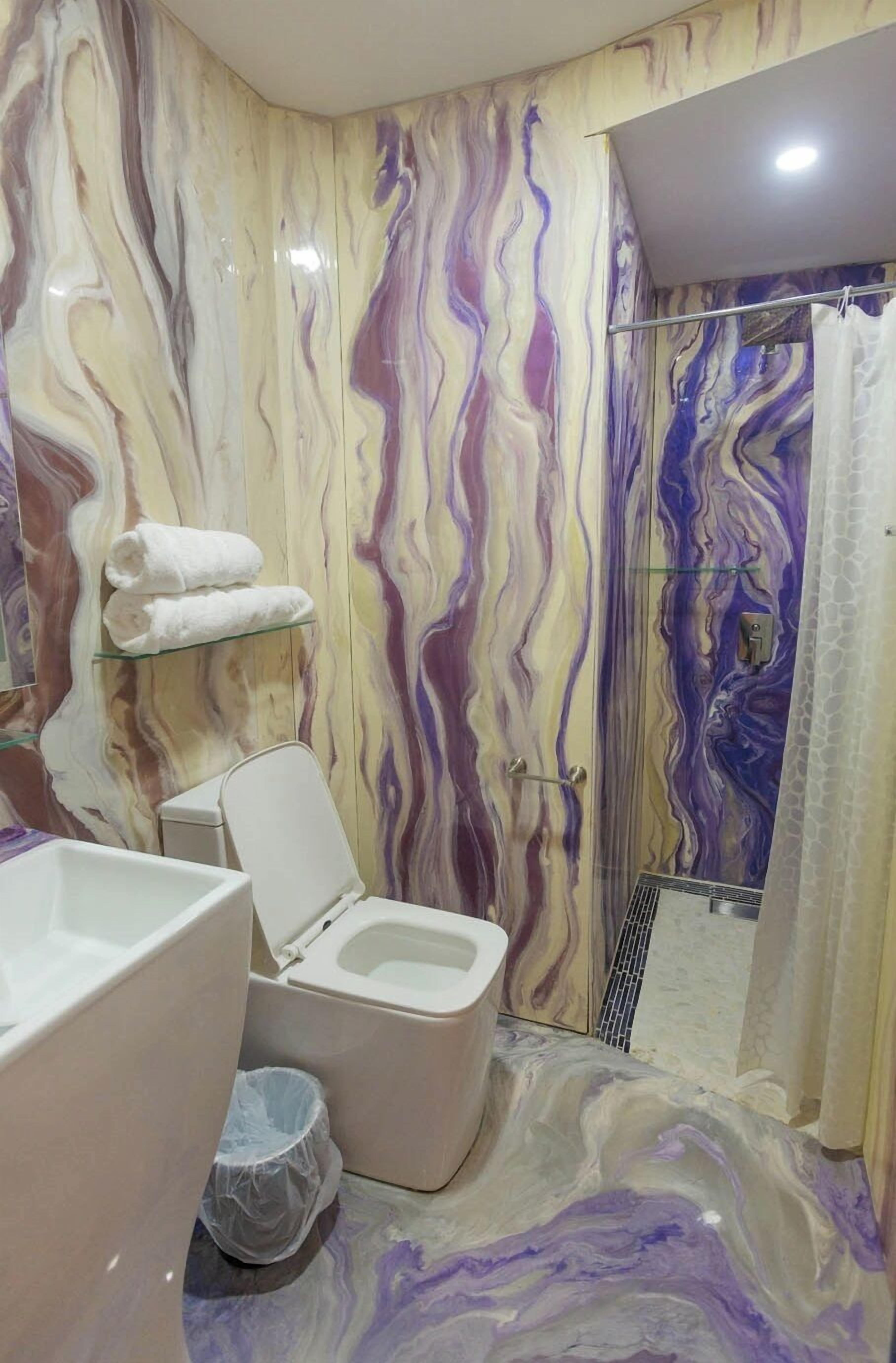 Un baño con pared de mármol, inodoro, ducha y repisa de vidrio con toallas.