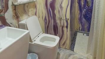 Un baño con pared de mármol, inodoro, ducha y repisa de vidrio con toallas.
