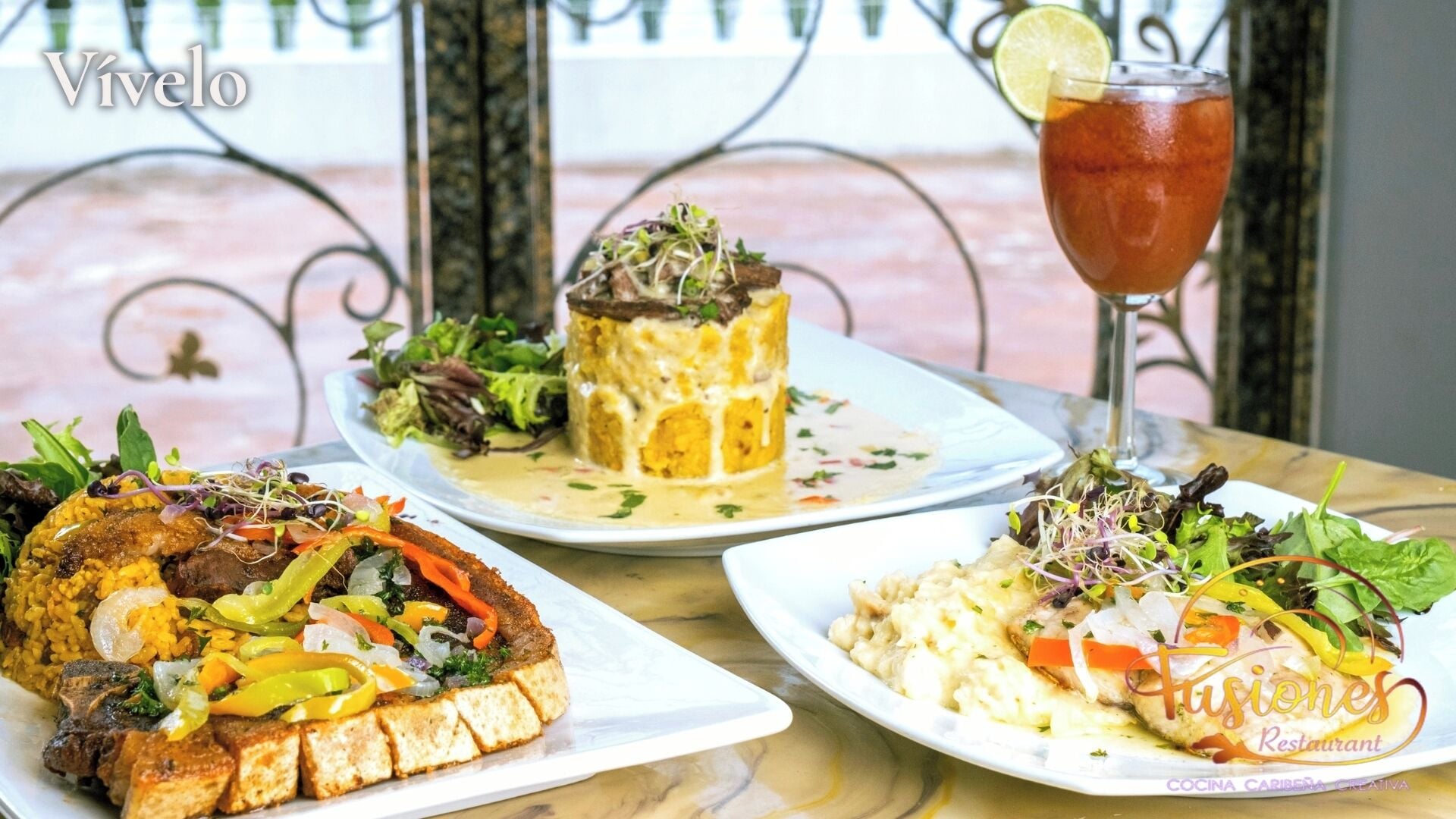 A table with three plates of food, a glass of iced tea, and a decorative metal railing in the background.