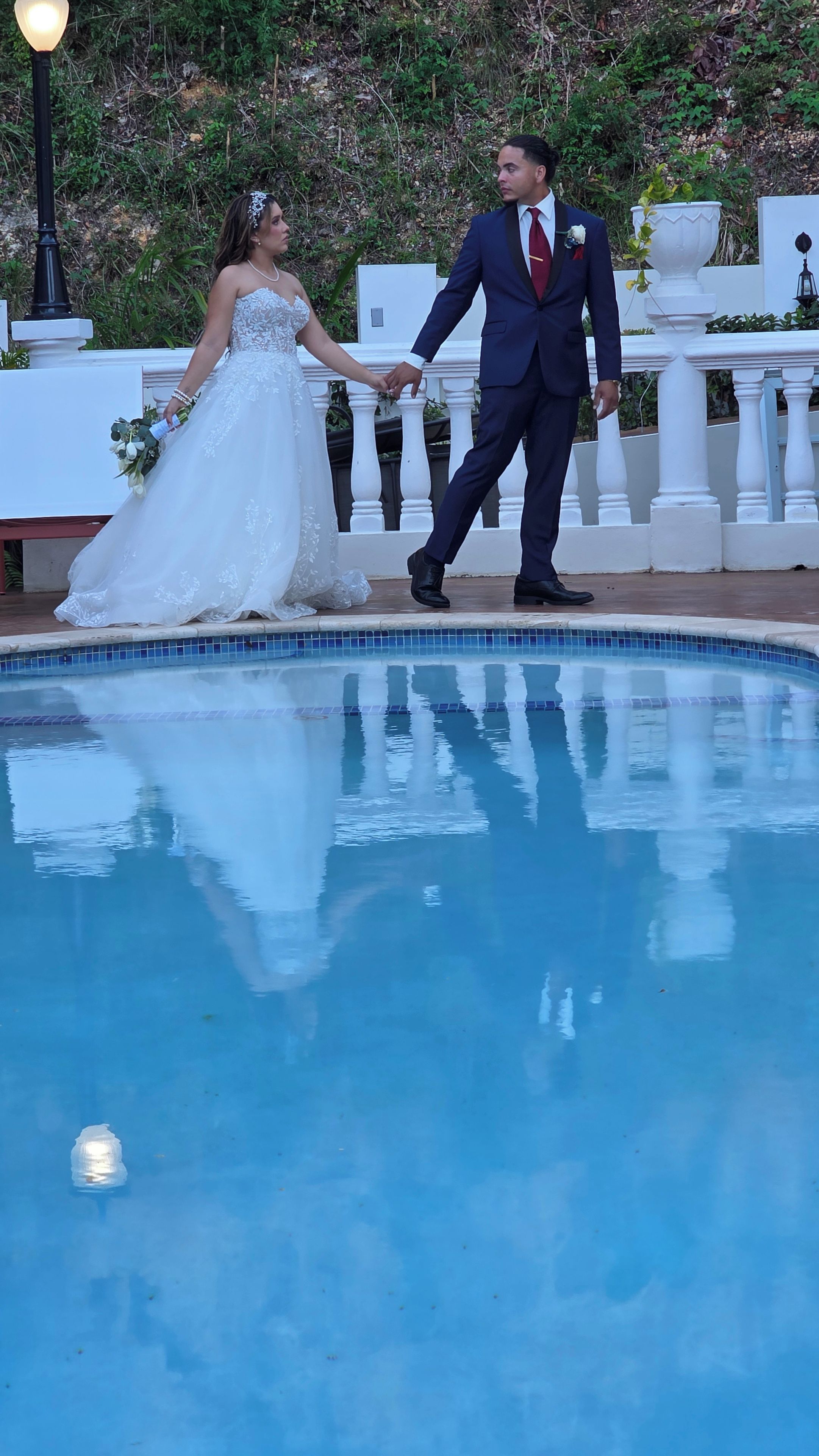 Un couple se tenant la main au bord d’une piscine, avec une balustrade blanche et de la verdure en arrière-plan.