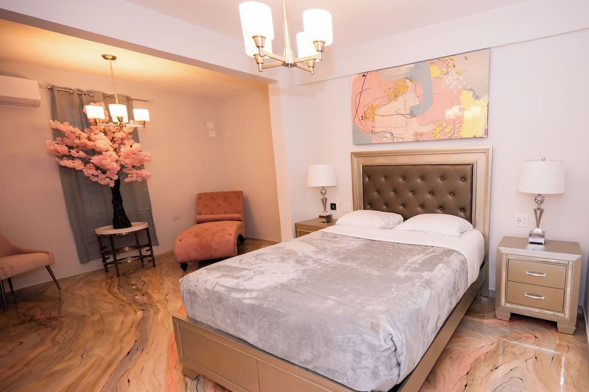A bedroom with a bed, a chair, a nightstand, a lamp, a vase with flowers, and a chandelier.