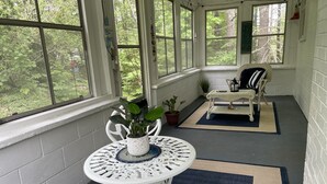 Une véranda couverte avec une table et des chaises blanches, des plantes en pot et une vue sur la verdure environnante.