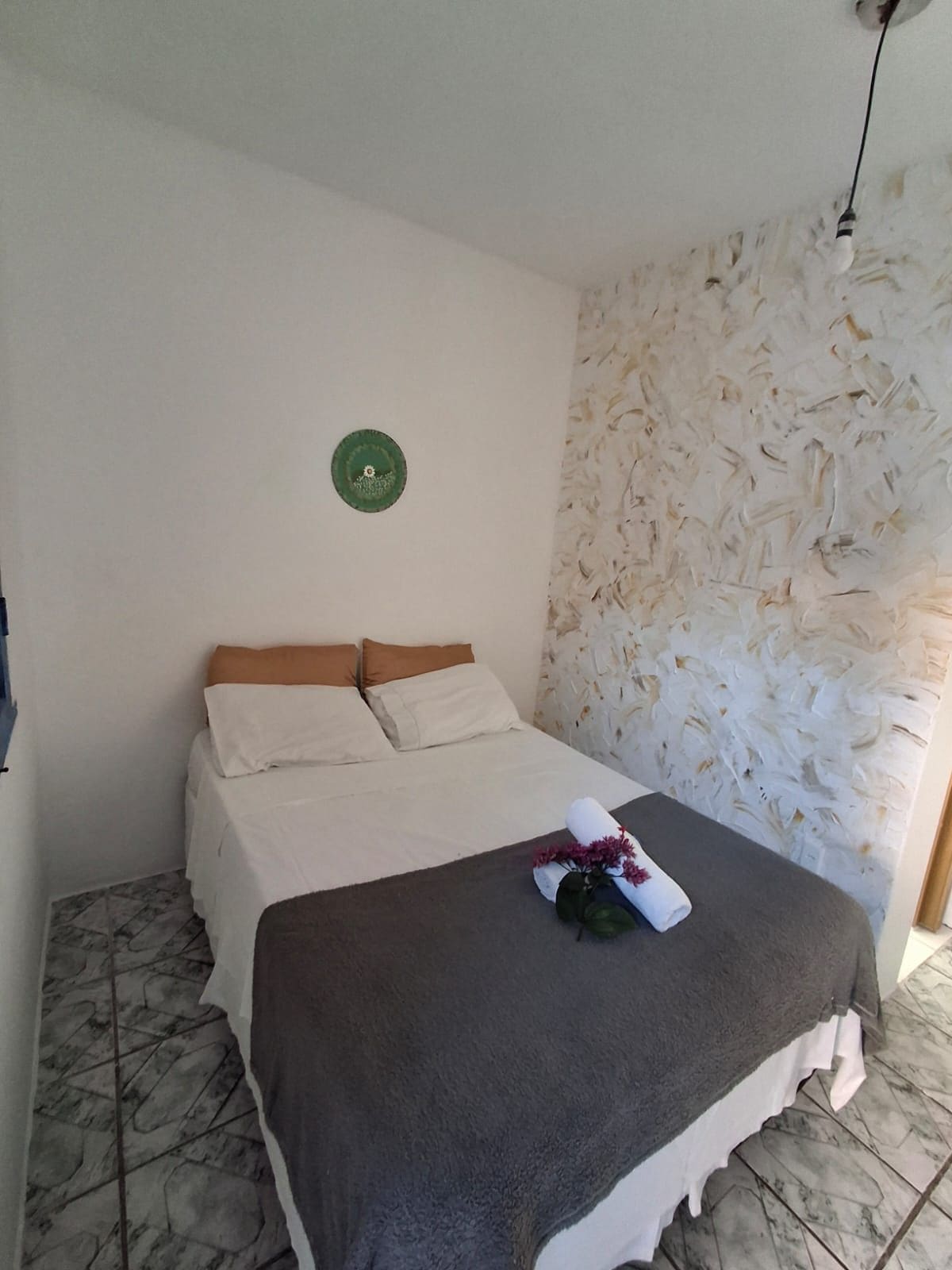 A single bed with a grey blanket and a rolled towel with flowers, a green wall decoration, and a textured wall to the right.