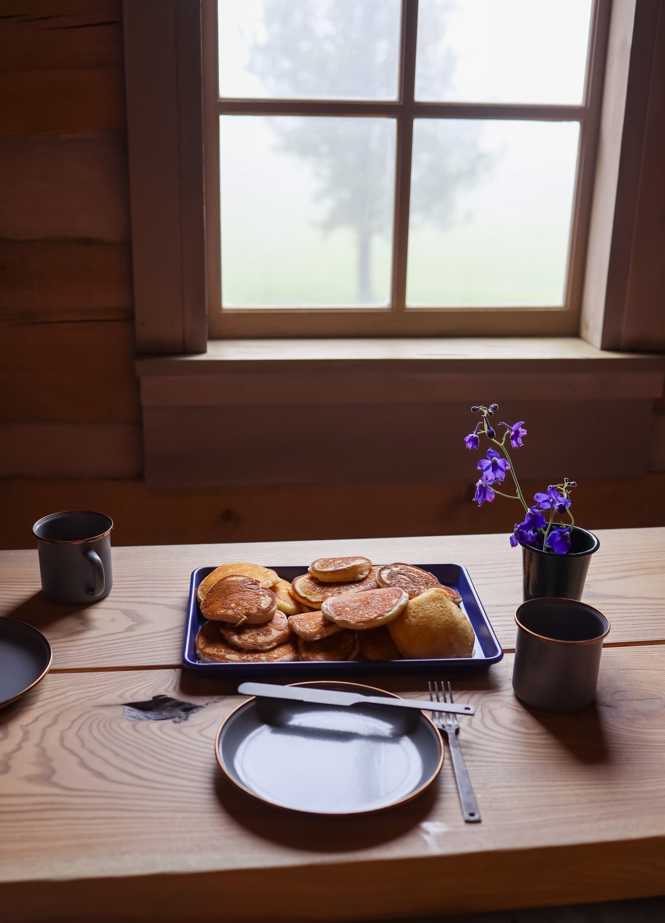 Une table en bois près d’une fenêtre, mise pour le petit-déjeuner, avec une assiette de crêpes, une assiette, une fourchette et une tasse.
