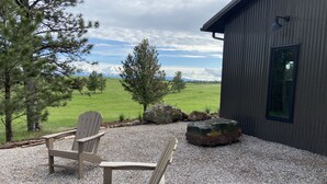 A patio with Adirondack chairs, a gravel ground, and a view of a green field and trees. - The Log House, Montana. Handmade & luxurious cabin. 10 min from Main St.  (Lewistown)