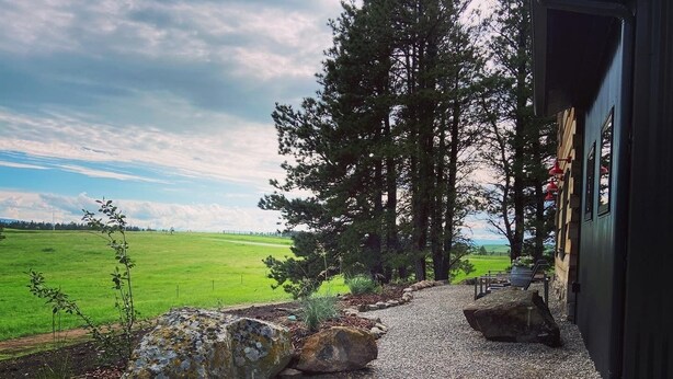 A gravel pathway leading to a building with a view of a green field and trees. - The Log House, Montana. Handmade & luxurious cabin. 10 min from Main St.  (Lewistown)