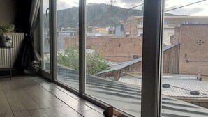 Ein Zimmer mit großen Fenstern, Holzboden und Blick auf eine Stadtlandschaft mit Gebäuden und einem Turm.