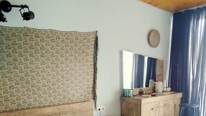 Ein Schlafzimmer mit einem Bett, Kissen, einer Holzkommode, einem Spiegel und einem Vorhang.