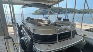 A stationary boat with a covered seating area, a name 'SunCruiser by Lowe' on the side, and a docked setting with a view of a lake and distant land. - Breathtaking Lake & Pool Views, In Osage Beach! (Osage Beach)