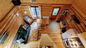 A wooden cabin interior with a sofa, dining table, kitchen area, and a window with curtains. - Chalet bois rond Prunier - Entièrement rénové! (Rivière-Rouge)