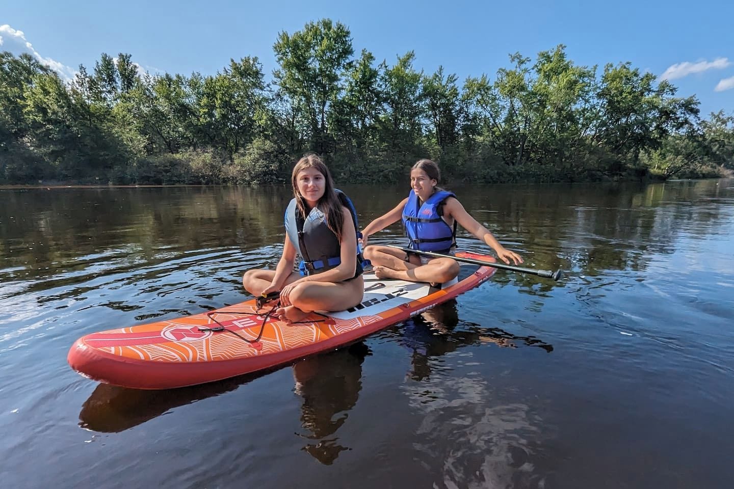 Two individuals sitting on a paddleboard in a calm body of water, surrounded by trees.