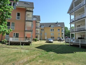 Un immeuble résidentiel à plusieurs étages, doté de balcons, d’une cour verdoyante et de voitures garées.