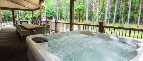 A hot tub in a covered outdoor area with a wooden deck, wicker furniture, and a view of a forest.