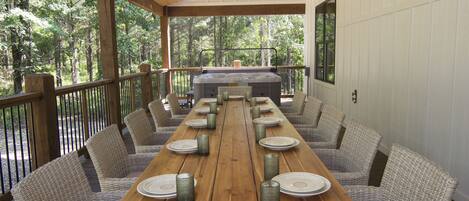 A long wooden table set for dining with wicker chairs, a hot tub, and a view of the forest.