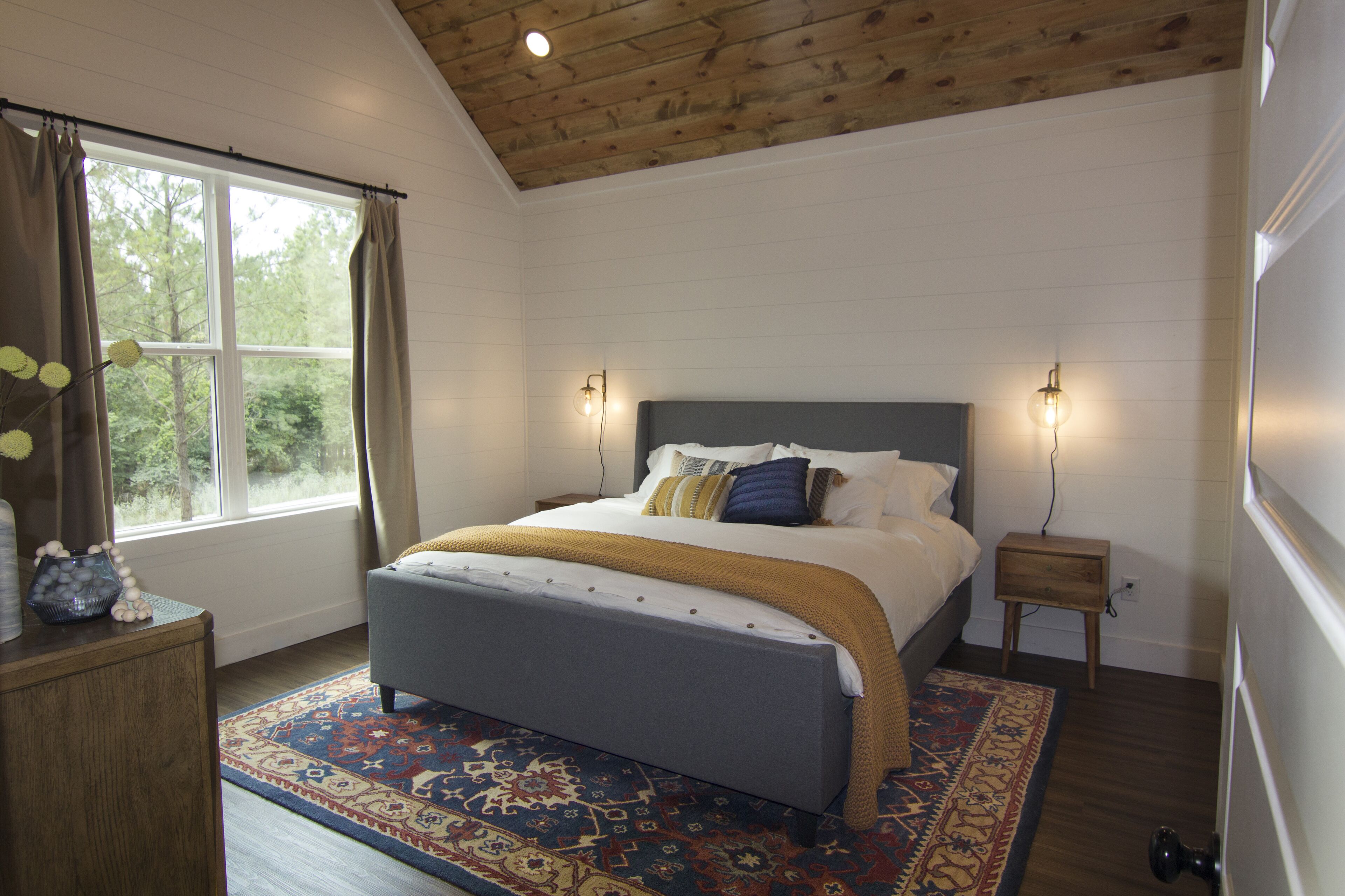Een slaapkamer met een bed, nachtkastje, dressoir en een raam met gordijnen.