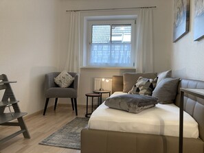 Ein Schlafzimmer mit einem Bett, einem Sessel, einem kleinen Tisch und einem Fenster mit Vorhängen.