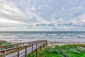 Ein hölzerner Steg führt zu einem Strand, wo Wellen an die Küste brechen, und der Himmel ist bewölkt.