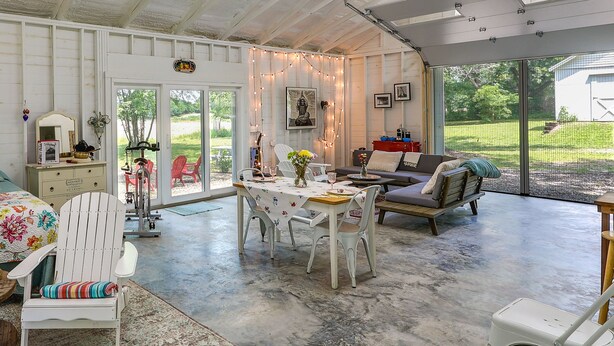 A cozy indoor space with a dining table, chairs, and a sofa. There is a white chair with a colorful cushion, a small table with a vase of flowers, and a sofa with cushions. The room has a concrete floor and white walls with a wooden ceiling. - Coffeyville Countryside Studio w/ Fire Pit! (Coffeyville)