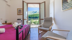 Une chambre avec un lit, une chaise, une petite table, un radiateur et un balcon donnant sur un paysage verdoyant.