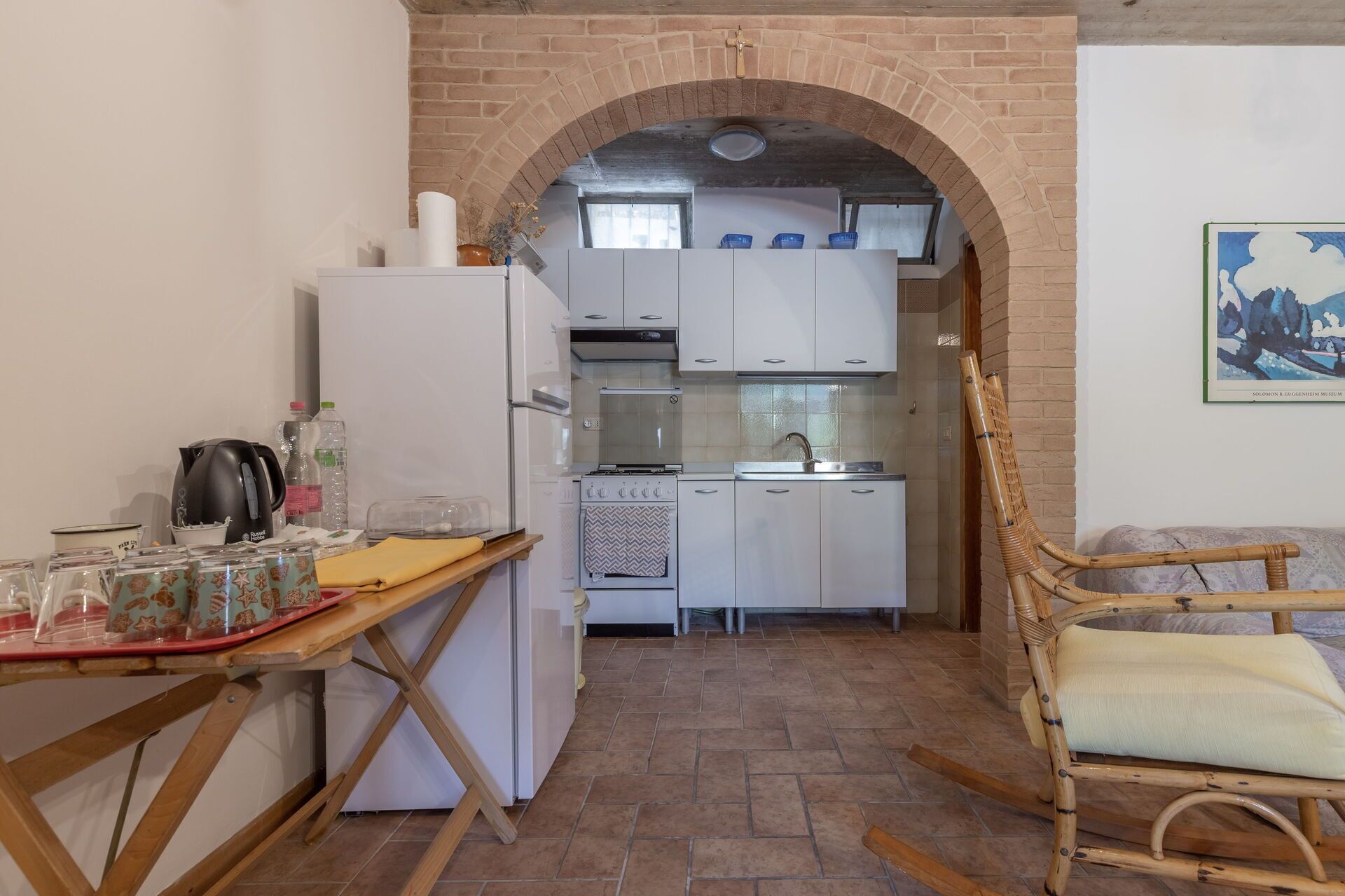 Une cuisine compacte avec des appareils électroménagers blancs, une table en bois sur laquelle sont posés des ustensiles de cuisine, et un arc en briques au style rustique.