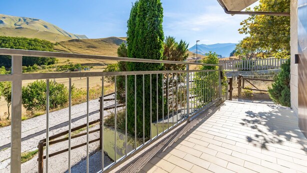 Un balcon doté d’une balustrade en métal, qui offre une vue sur un paysage vallonné, avec un grand arbre à feuilles persistantes et un ciel dégagé.