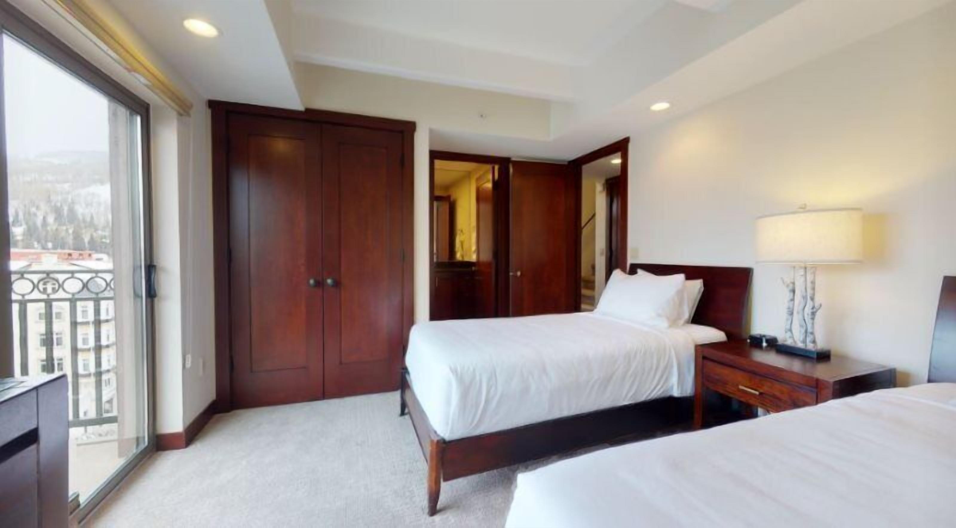 Ein Hotelzimmer mit zwei Betten, einem großen Holzschrank, einem Spiegel, einem Nachttisch mit Lampe und Blick auf einen Balkon.