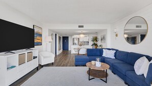 A modern living room with a large flat-screen TV, a blue sofa, a coffee table, and a dining area with a white table and chairs. - Anchorage Resort Siesta Key 2 Bedroom 6th Floor Condo Beach Access (Siesta Key)