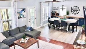 A modern living room with a sofa, coffee table, and dining area with chairs and a kitchen in the background. - Riverbend Ranch*Smith Lake Family Fun!*Dog Yard! (Crane Hill)