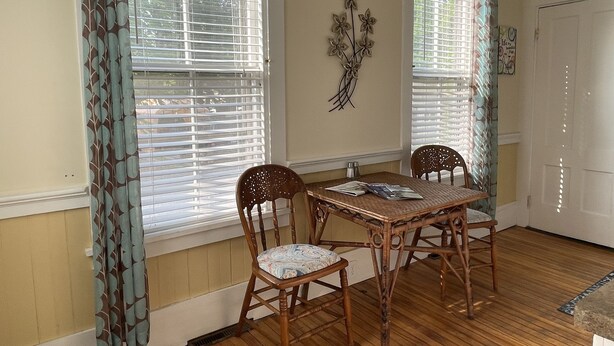 A dining area with a wooden table and chairs, large windows with blinds, and a decorative wall piece. - Cozy 1890 Cottage Studio with Hot Tub and Fire Pit in Downtown Kennebunk (Kennebunk)