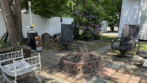 A patio with a fire pit, two wicker chairs, a metal bench, and a white fence. - Cozy 1890 Cottage Studio with Hot Tub and Fire Pit in Downtown Kennebunk (Kennebunk)