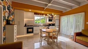 A modern kitchen with white cabinets, a dining area with a wooden table and chairs, and a living room with a sofa and curtains.